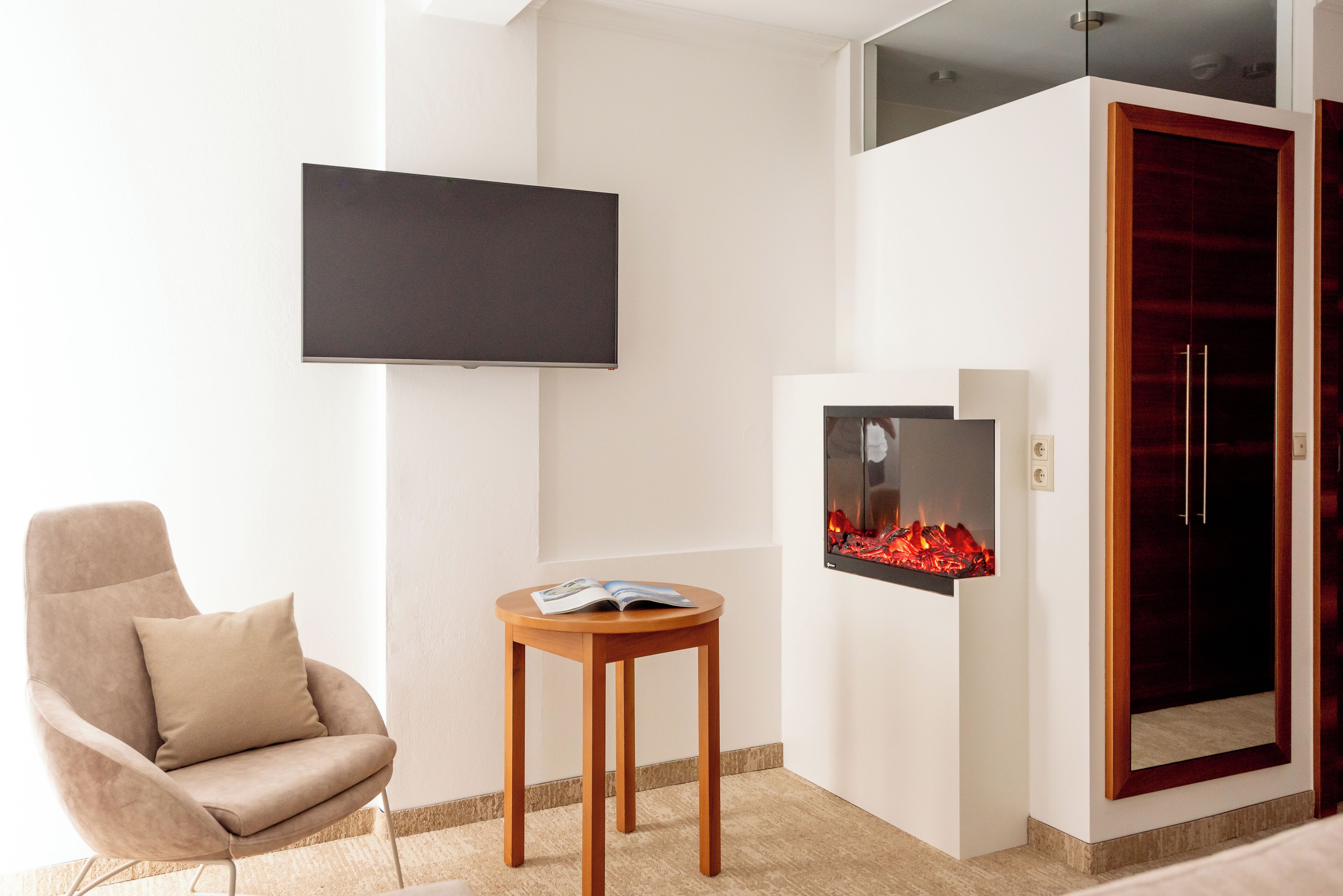 Una habitación moderna con un televisor de pantalla plana montado en la pared, un sillón cómodo, una pequeña mesa de madera con una revista y una chimenea empotrada.
