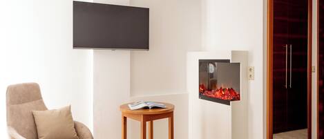 Una habitación moderna con un televisor de pantalla plana montado en la pared, un sillón cómodo, una pequeña mesa de madera con una revista y una chimenea empotrada.