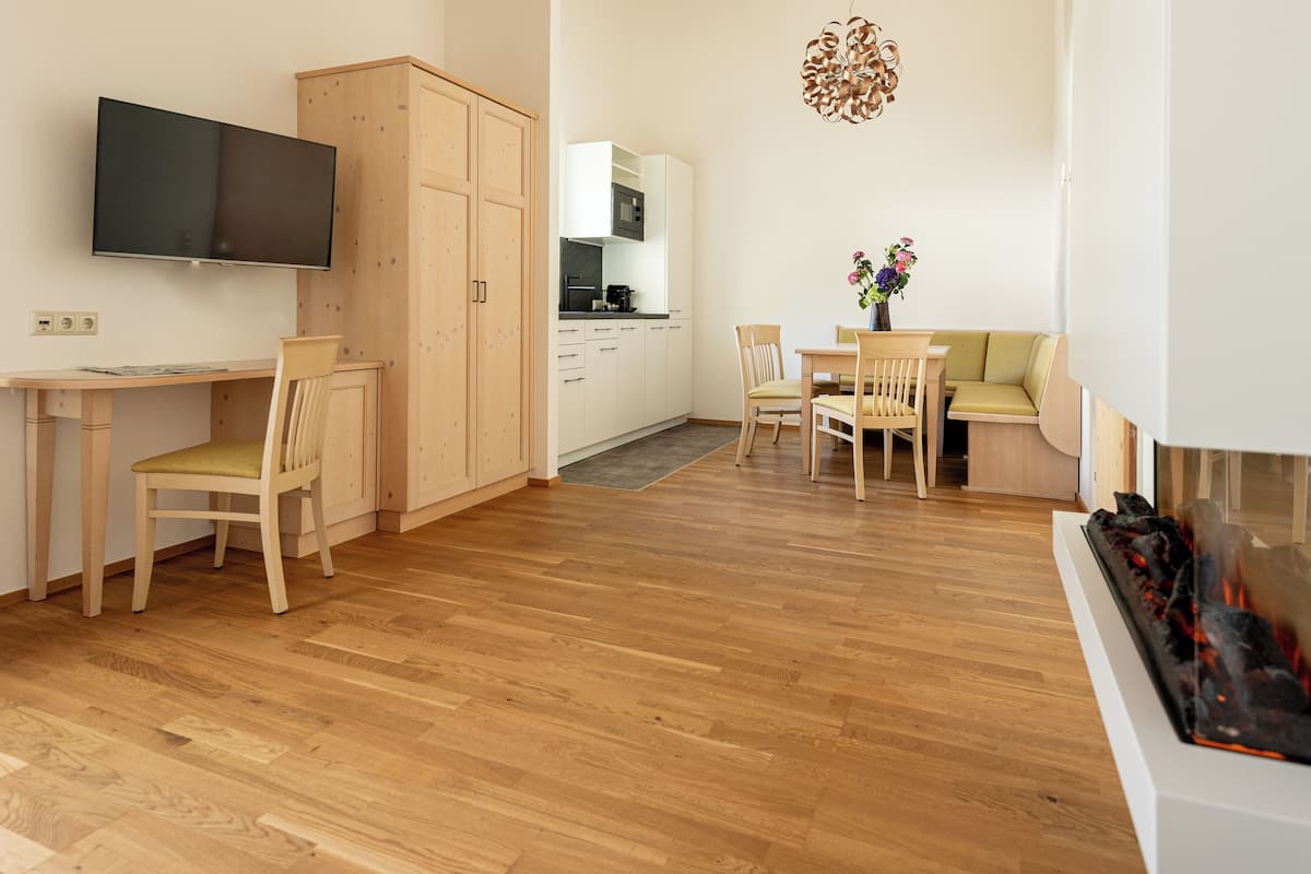 A modern, open-plan living and dining area with wooden flooring, a flat-screen TV mounted on the wall, a dining table with chairs, and a kitchen area with white cabinets and a microwave.