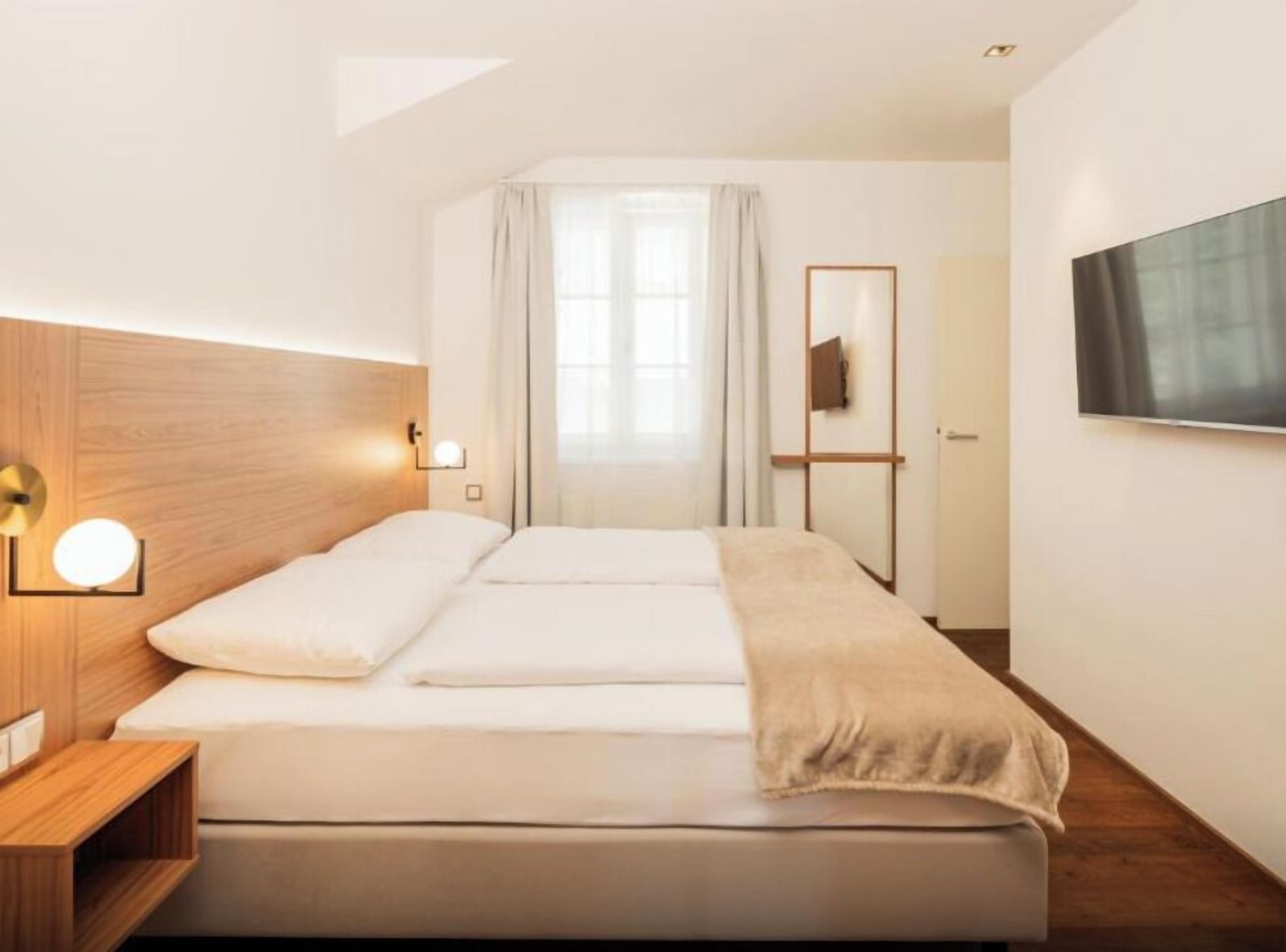 Una habitación de hotel moderna con cabecera de madera, cama con ropa de cama blanca, mesita de noche con lámpara, ventana grande con cortinas y televisor de pantalla plana montado en la pared.