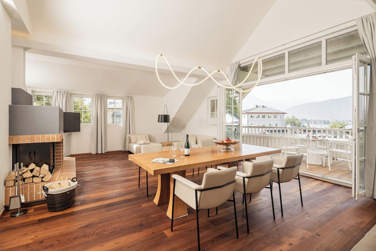 A spacious dining area with a long wooden table, several chairs, a fireplace, and a large window offering a view of the city.