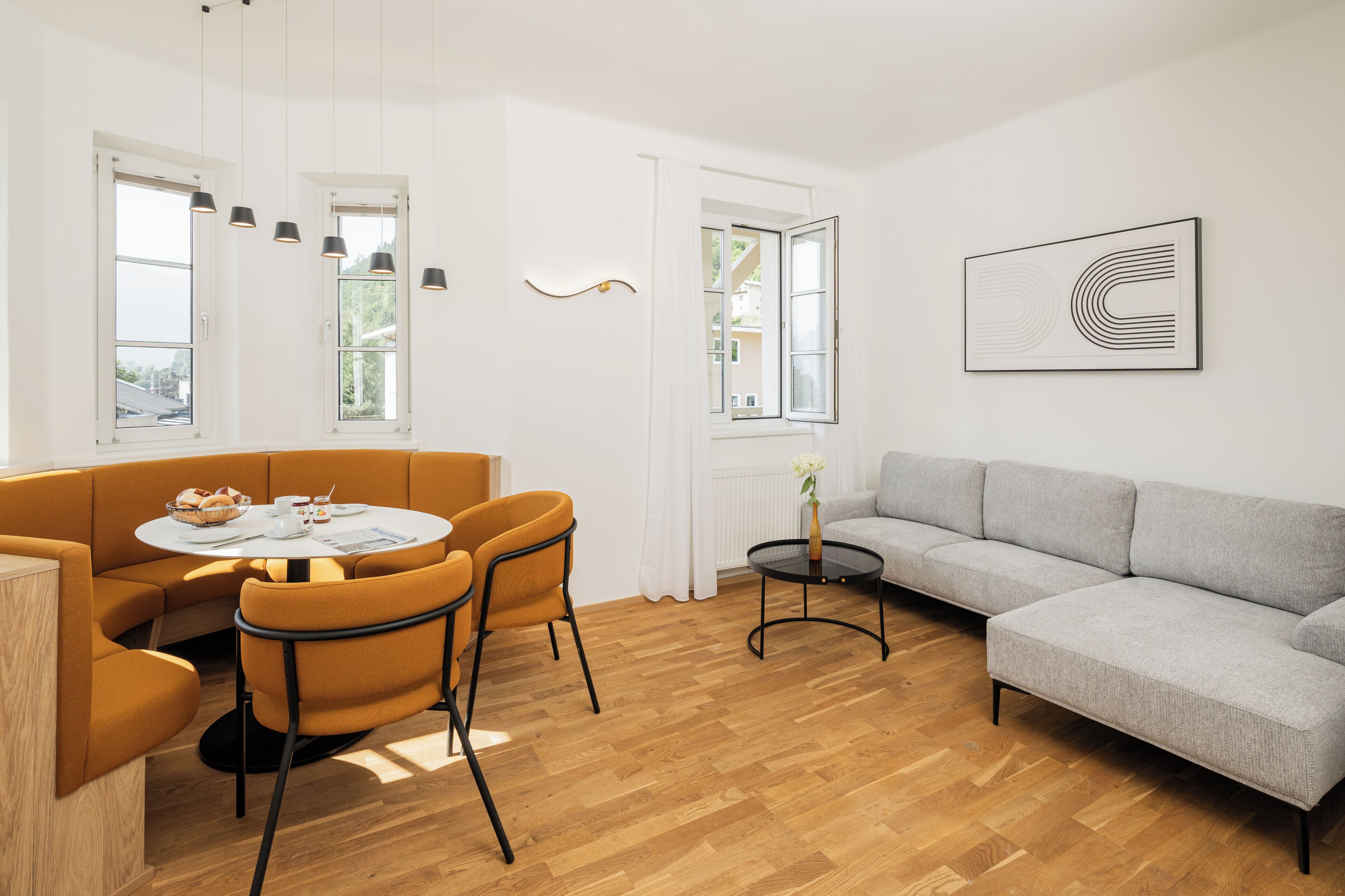 Ein modernes Wohnzimmer mit einer Couch, einem runden Tisch und Stühlen. Es gibt Fenster und ein Wandbild.