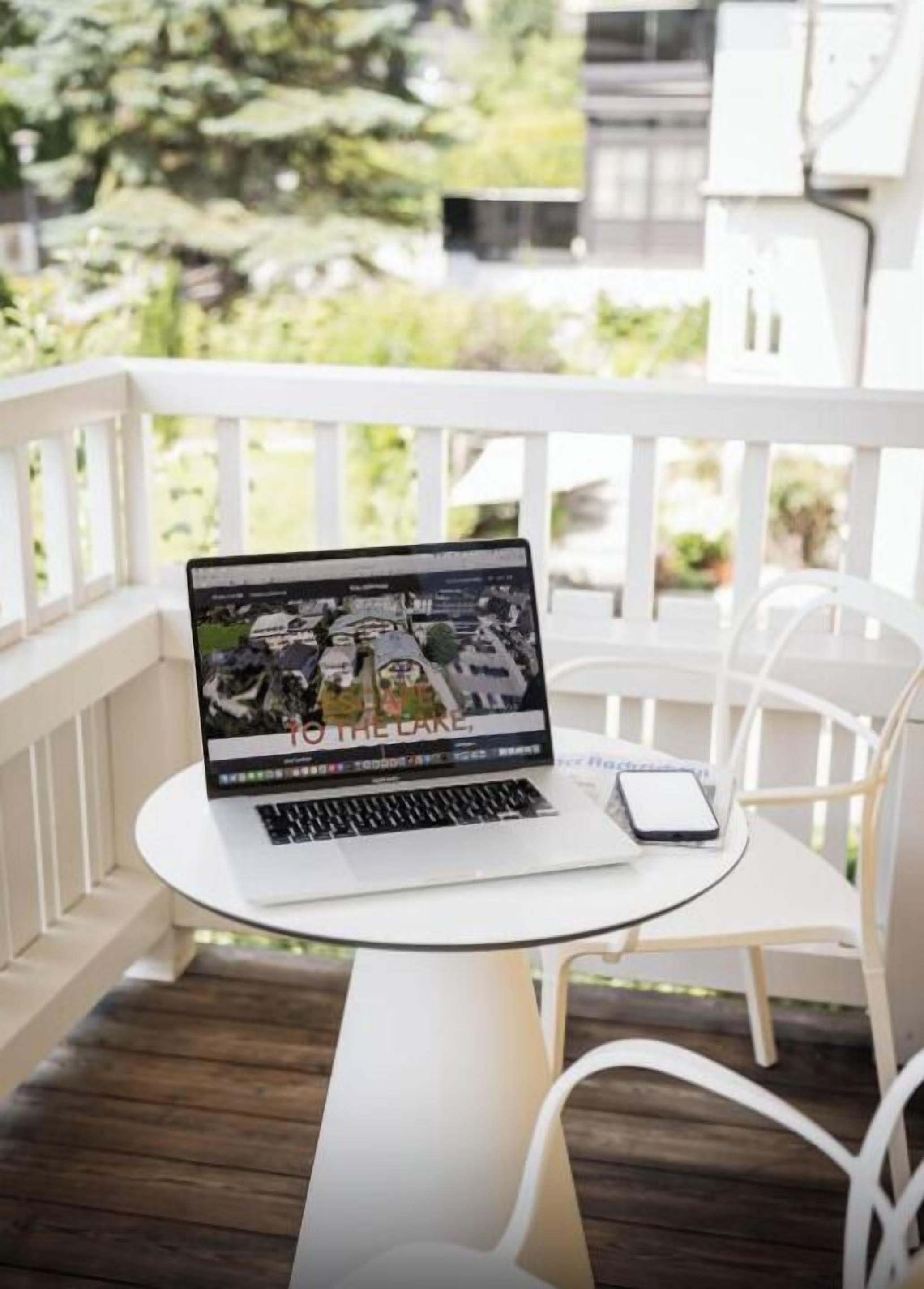 A laptop on a white table on a balcony with white chairs, a white railing, and a view of greenery.
