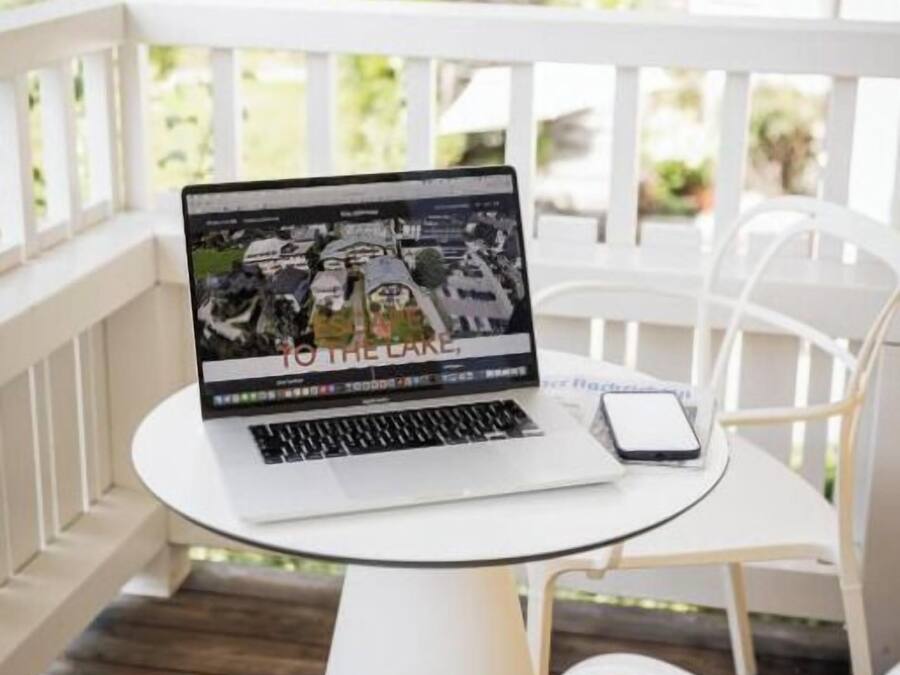 A laptop on a white table on a balcony with white chairs, a white railing, and a view of greenery.