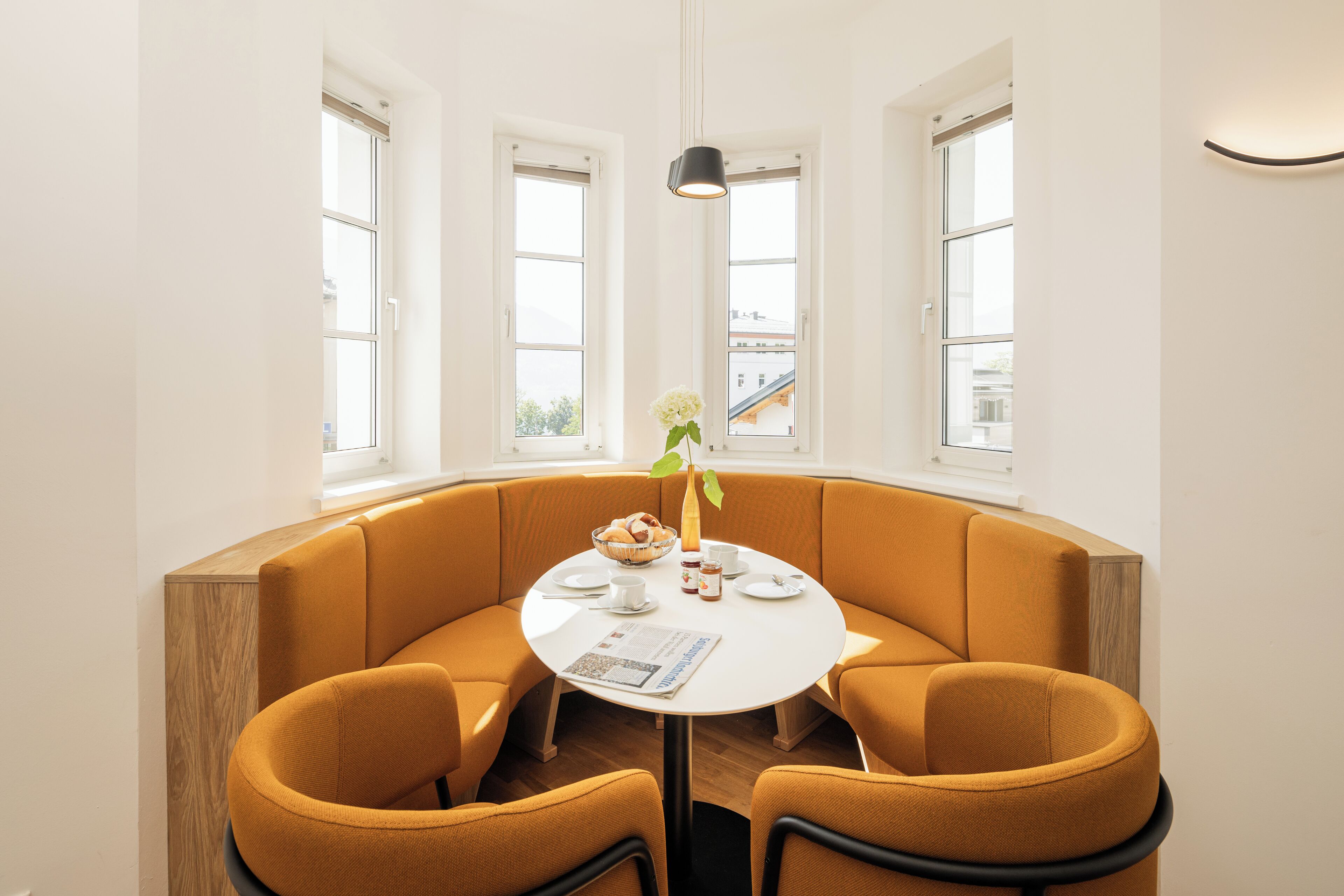 A corner dining area with a white table and orange upholstered chairs, a vase with flowers, and a window with a view of buildings.