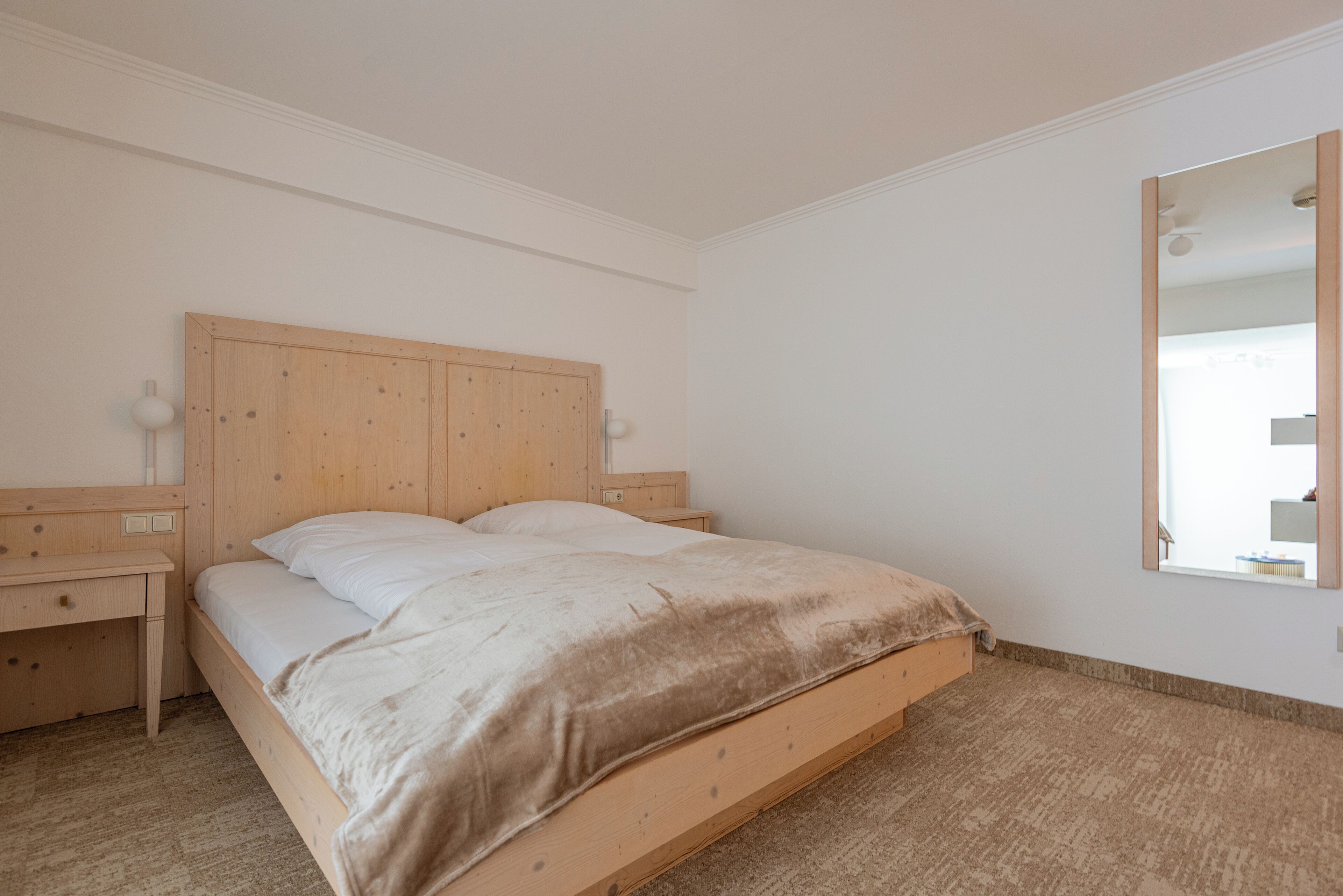 Un dormitorio con una cama de madera, dos mesitas de noche, un buró, un espejo grande y piso alfombrado.