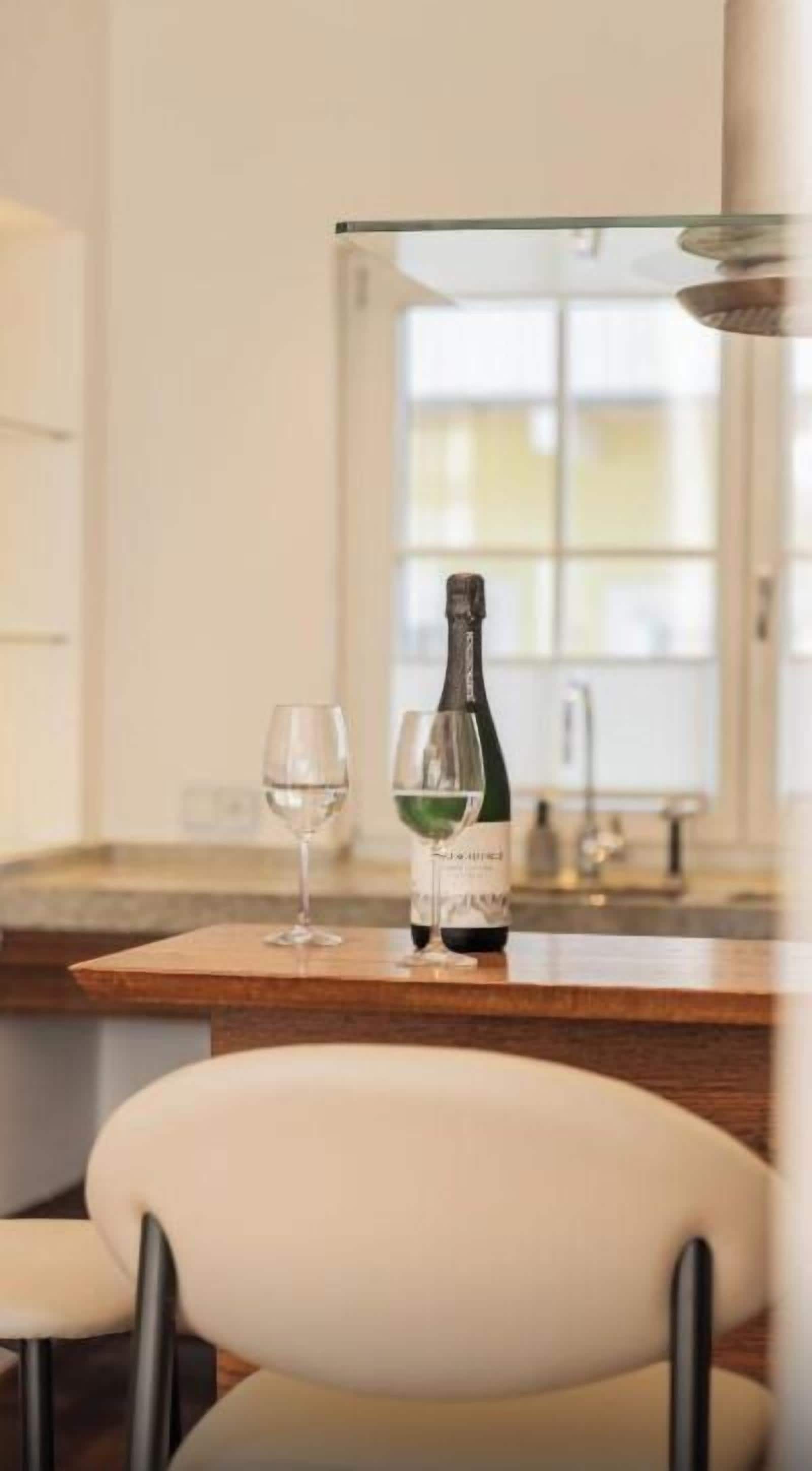 A bottle of wine and a glass on a wooden table in a well-lit room with a window in the background.