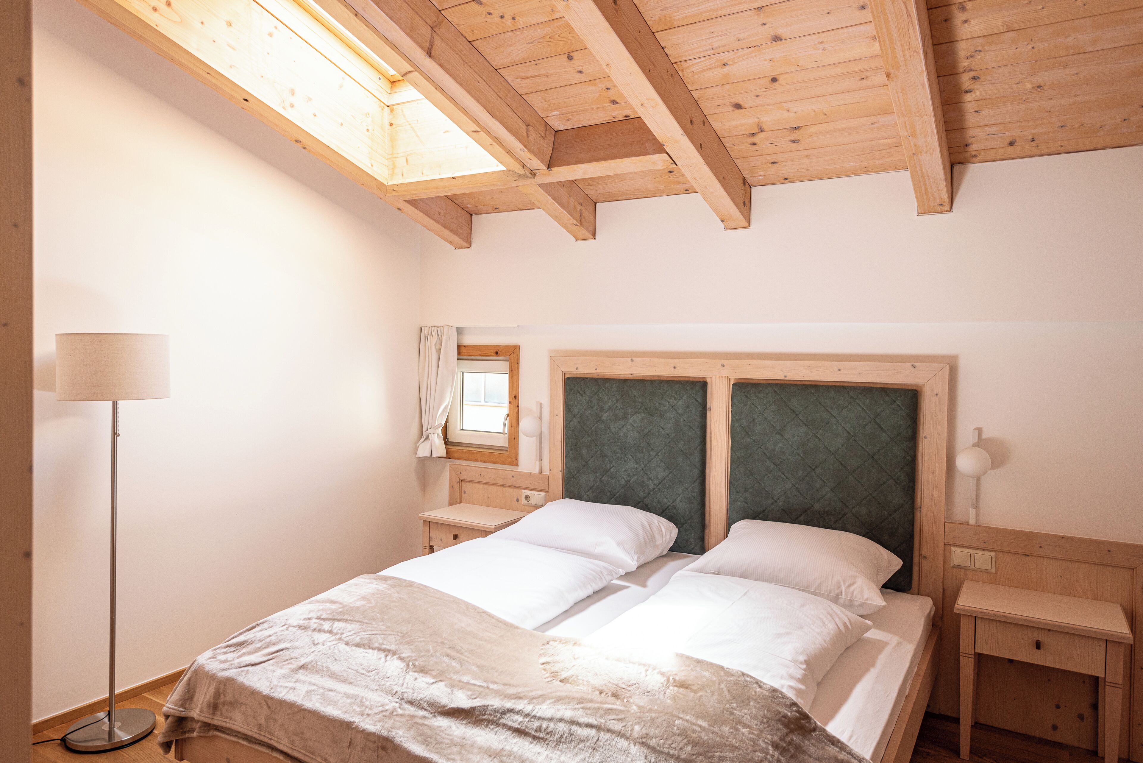 Un dormitorio con techo de madera, una cama con ropa blanca, una mesita de noche y una lámpara de pie.