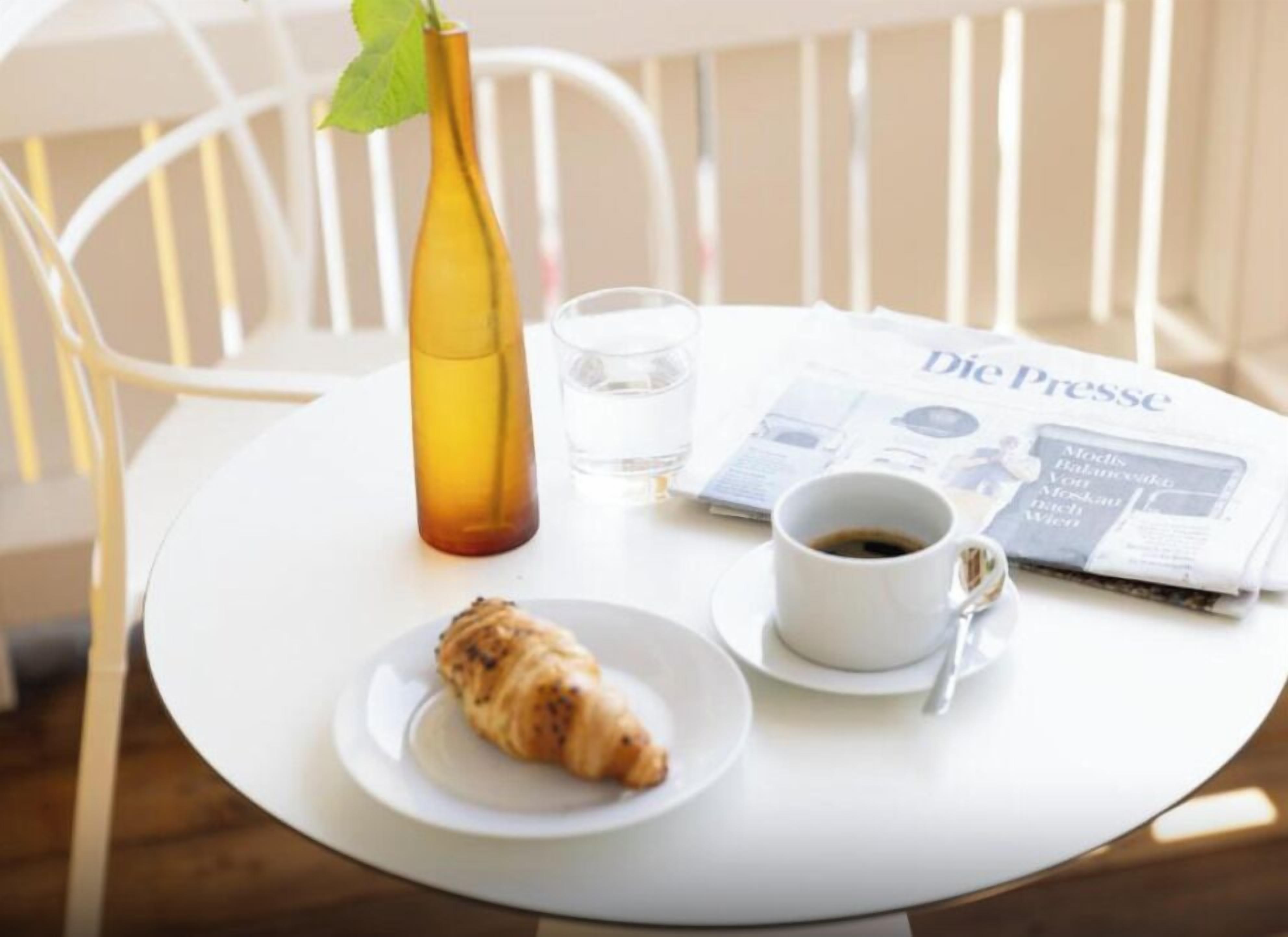 Una mesa con un croissant, una taza de café, un vaso de agua, un periódico y una botella amarilla.