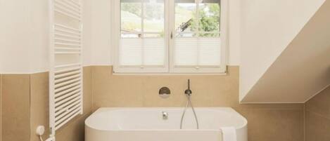 A modern bathroom with a freestanding bathtub, a window with shutters, and a towel hanging on the tub.