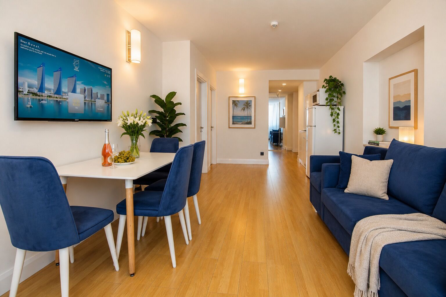 Una sala de estar moderna con un sofá azul, una zona de comedor con una mesa blanca y sillas azules, un televisor montado en la pared y un pasillo que conduce a la cocina.