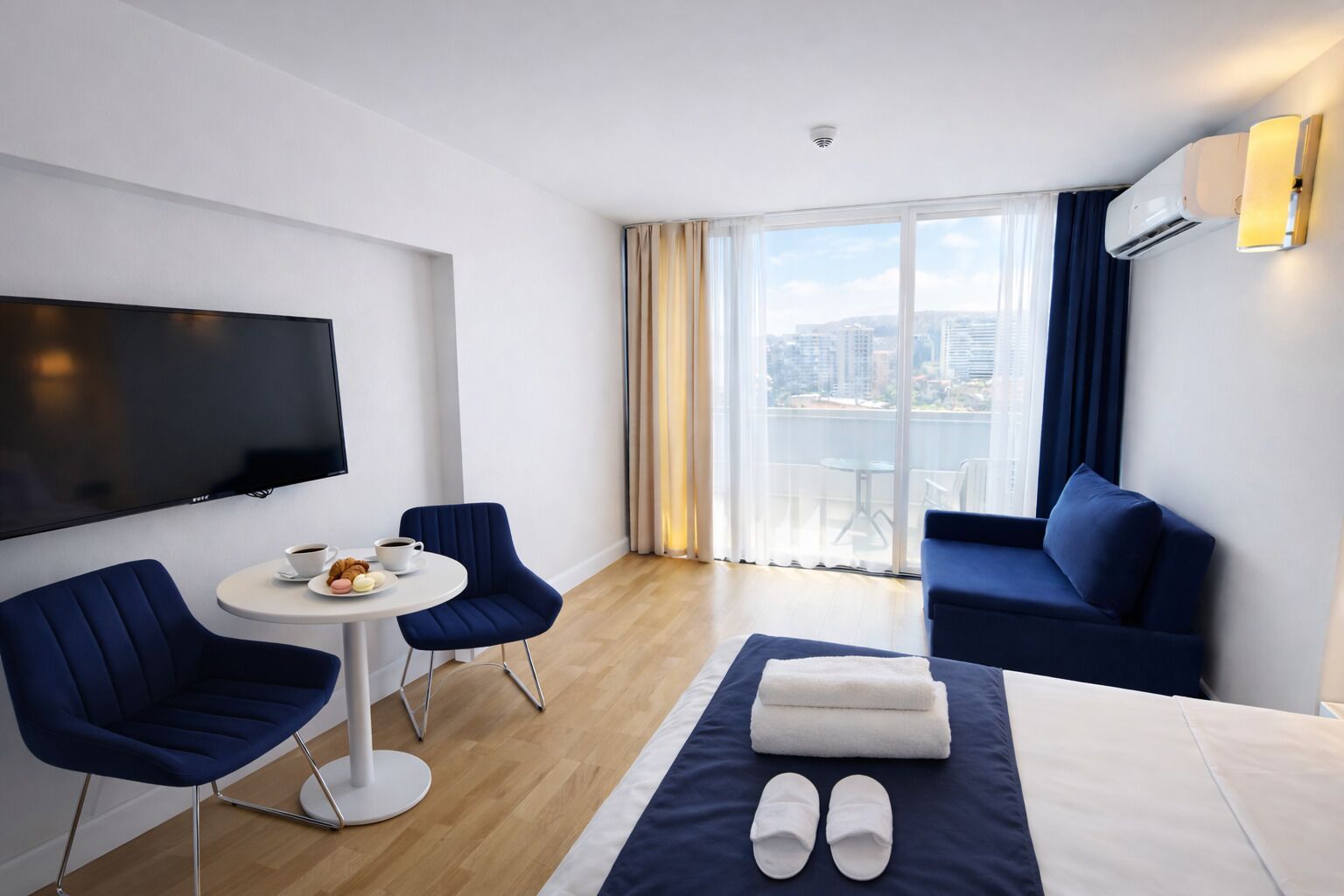 A hotel room with a bed, a TV, two chairs, a small table with breakfast, a sofa, and a balcony with a city view.