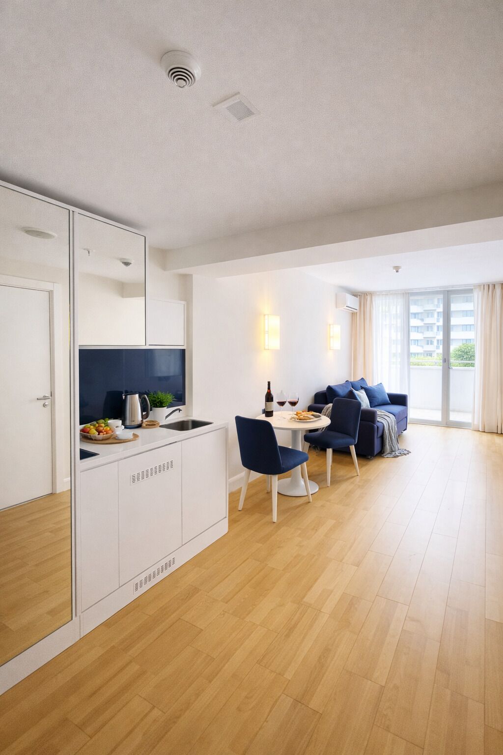 A modern kitchen with white cabinets, a dining area with a round table and blue chairs, and a living area with a sofa and a view of the outside.