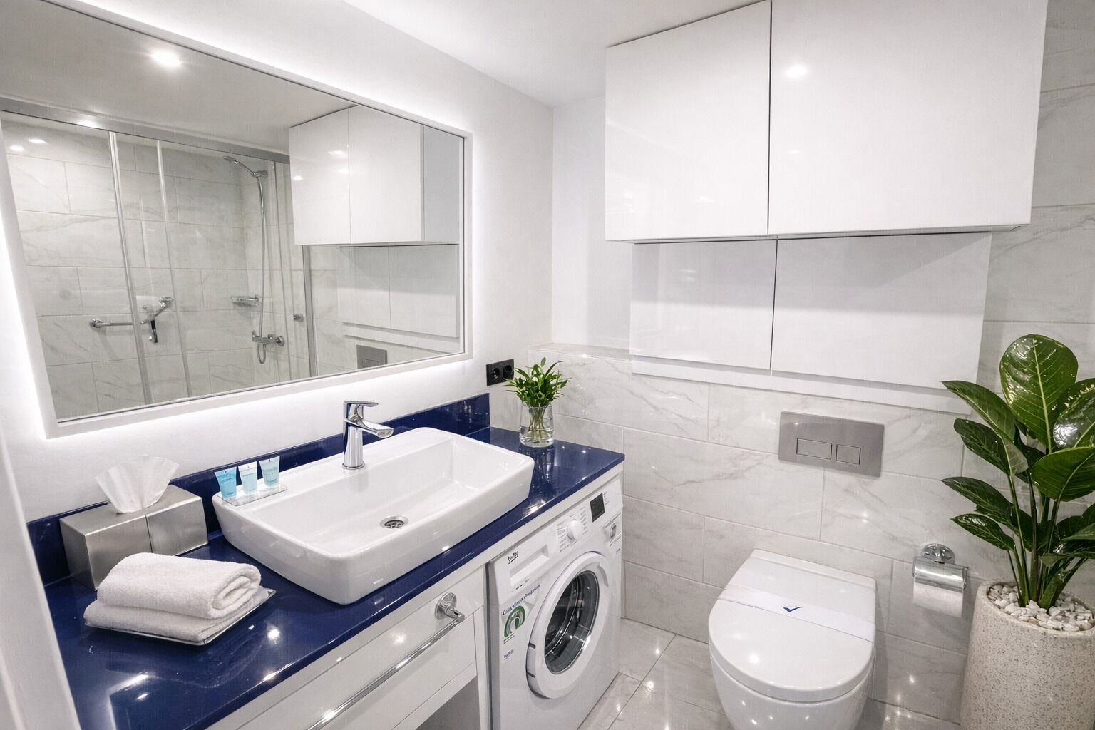 A modern bathroom with a large mirror, a white sink, a washing machine, and a toilet.