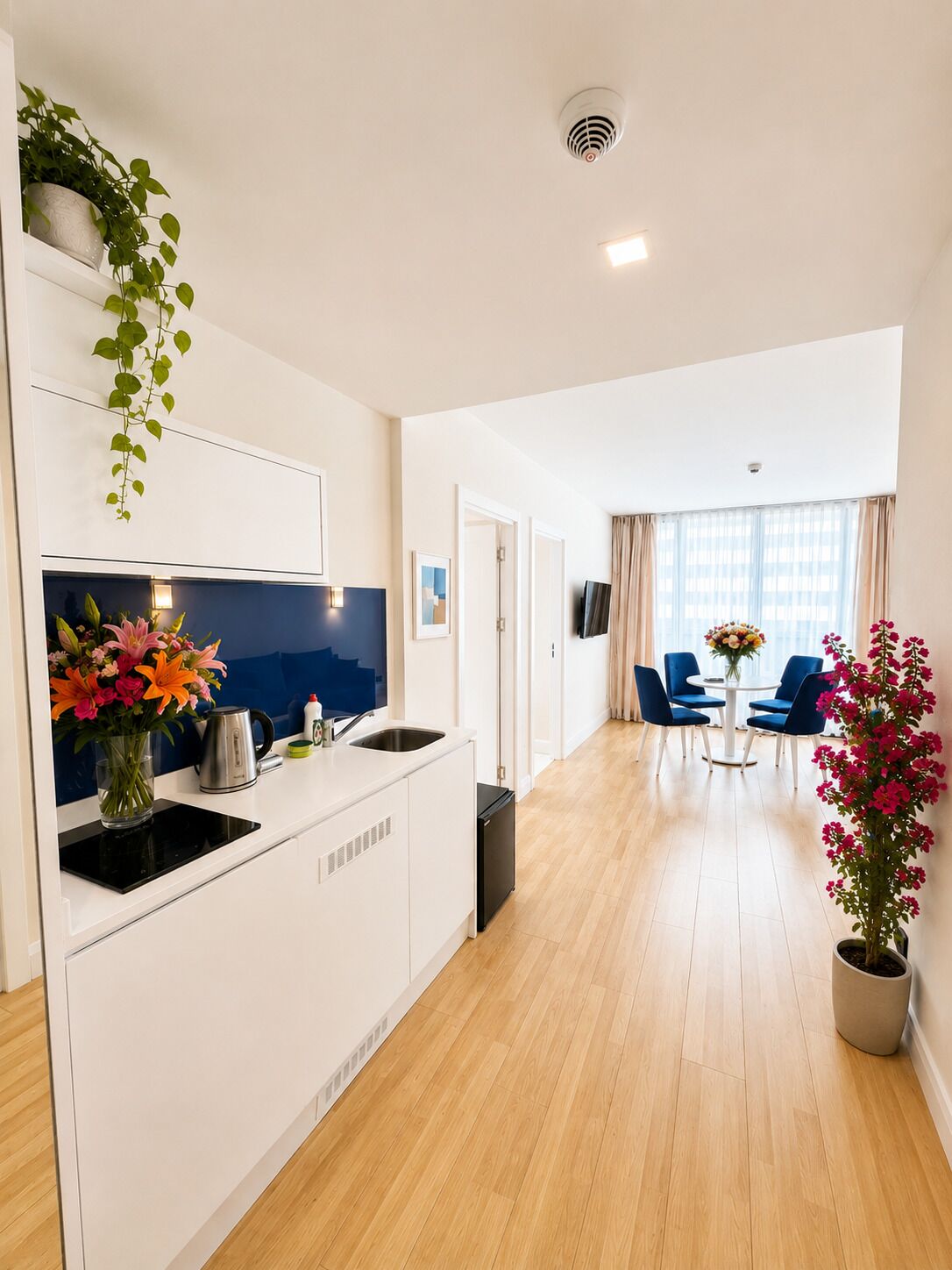 Uma cozinha moderna com forno embutido, pia e cafeteira. Há uma área de refeições com cadeiras azuis e uma mesa, e uma sala de estar com TV e planta.