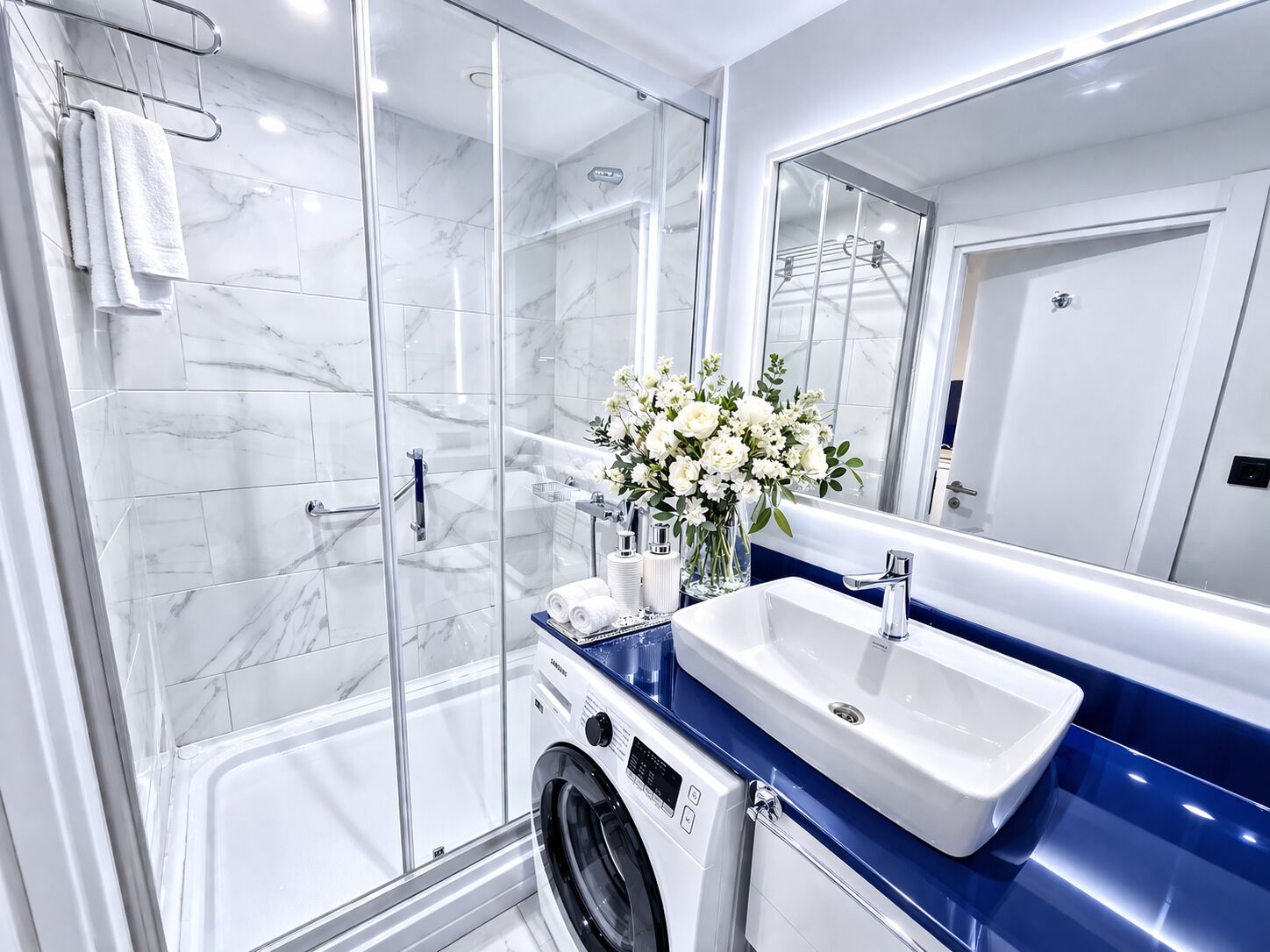 A modern bathroom with a glass shower enclosure, a built-in washing machine, a sink, and a mirror.