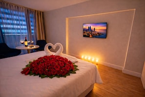 Une chambre d’hôtel avec un lit, un bouquet de roses rouges, une décoration en forme de cœur réalisée avec des serviettes, un tableau représentant un paysage urbain, et une table avec une bouteille de vin et des verres.