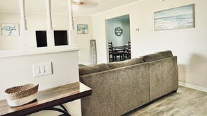 Un salon avec un canapé gris, une table en bois sur laquelle se trouve un panier en osier, un coin repas avec une horloge et un tableau accroché au mur.