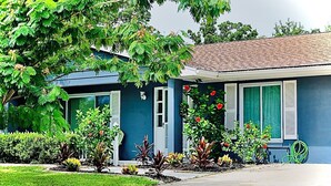 Une maison d’habitation à l’extérieur bleu, avec un jardin devant la maison où l’on peut voir différentes plantes et fleurs, et une porte de garage bien visible.