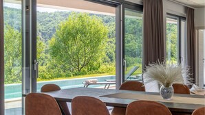 A dining area with a table, chairs, and a view of a swimming pool and forested hills.