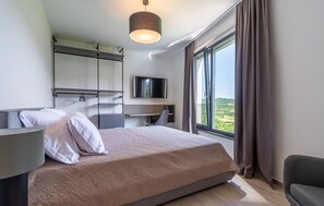 Une chambre d’hôtel moderne équipée d’un lit, d’un bureau, d’une télévision et d’une grande fenêtre donnant sur la campagne.