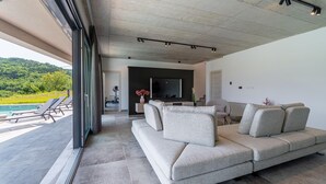 A modern living room with a grey sectional sofa, a flat-screen TV mounted on the wall, and a pool visible through the sliding glass doors.