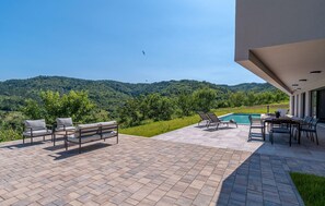 Une terrasse avec mobilier d’extérieur, une piscine et une vue sur une colline boisée.
