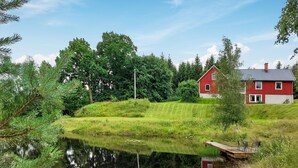 Ein rotes Haus mit geneigtem Dach, umgeben von üppiger Vegetation und einem stillen Teich.