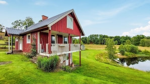 Ein rotes Holzhaus mit Veranda, umgeben von einer üppigen grünen Landschaft und einem Gewässer.