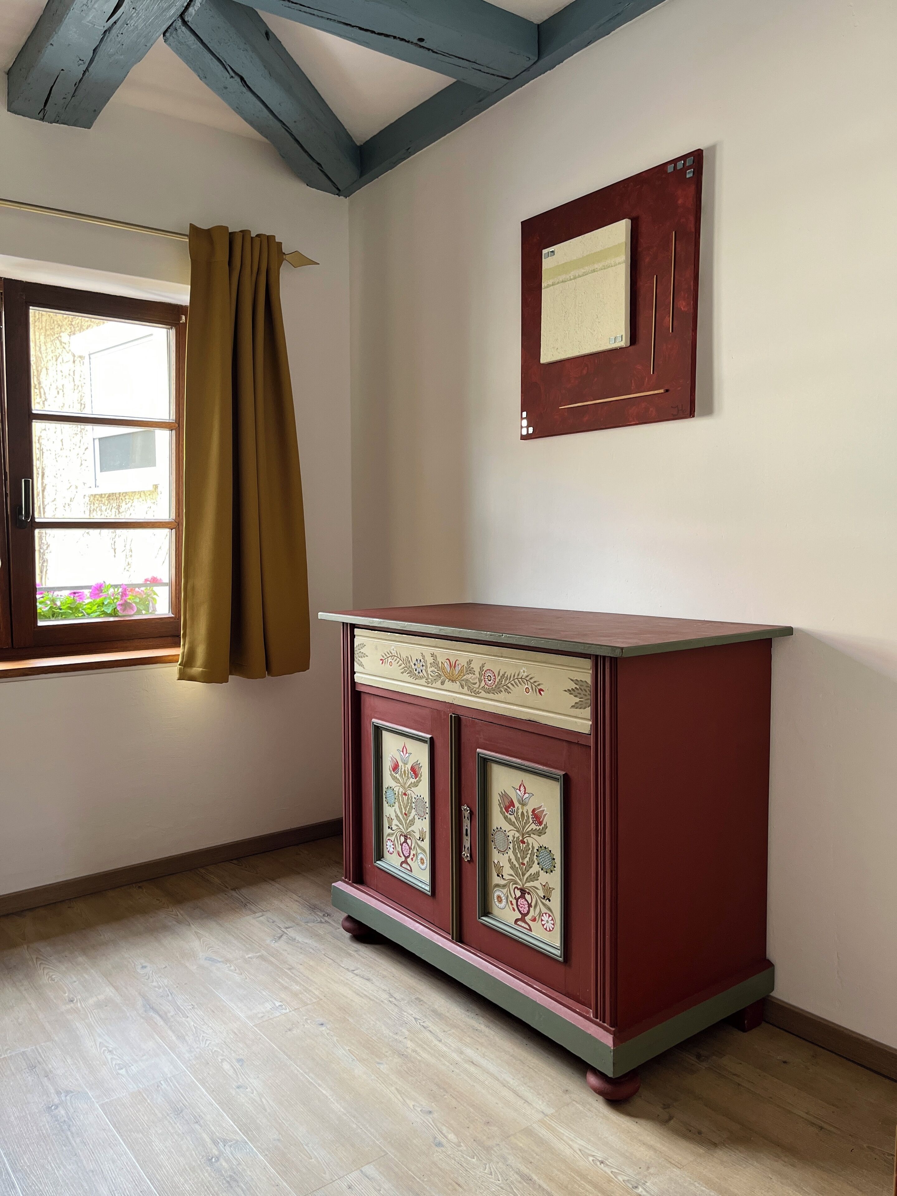 Una stanza con pavimento in legno, un armadietto rosso decorato con motivi floreali, una finestra con tenda marrone e un quadro appeso al muro.