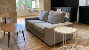 A living room with a grey sofa, a round wooden table, and a white side table. There is a black kitchen area with a stove and cabinets. - Bezenac grocery store (Castels et Bézenac)
