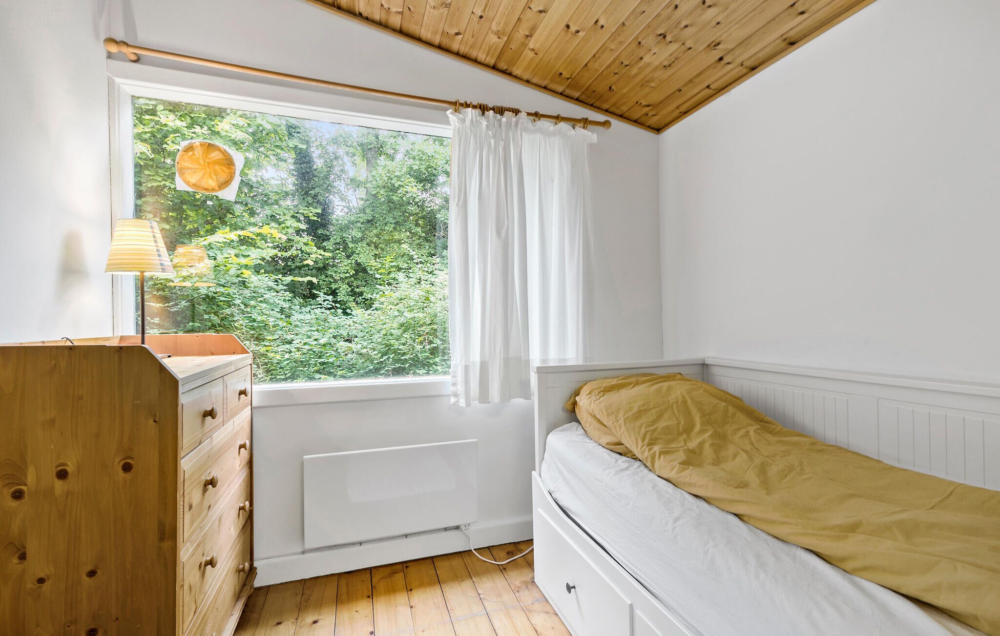 Un dormitorio con techo de madera, una ventana con cortinas blancas, una cama con manta amarilla, un aparador de madera y un radiador.
