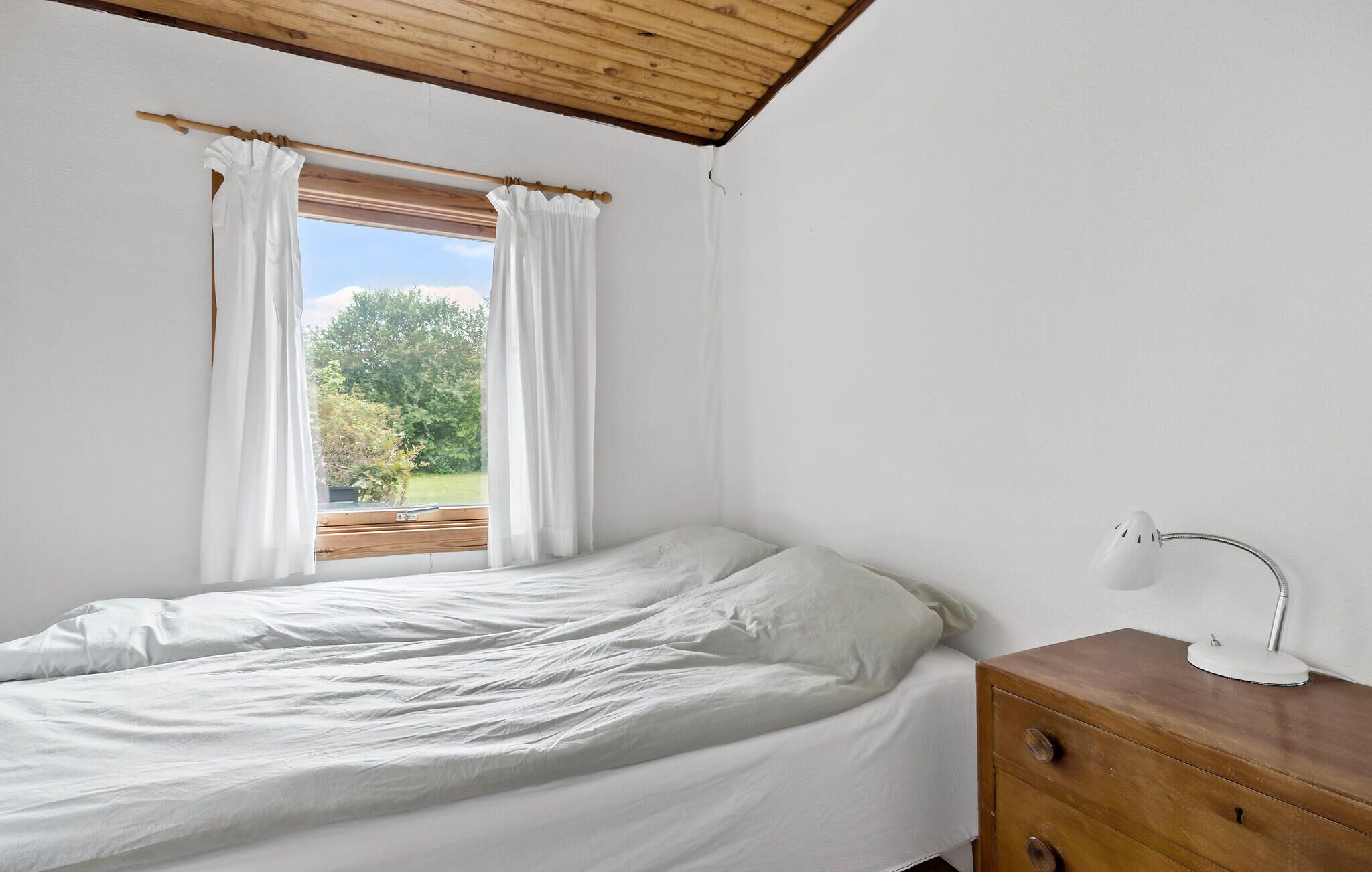Een slaapkamer met een bed, een houten dressoir, een bureaulamp en een raam met gordijnen.