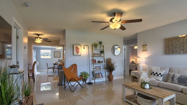 A spacious living room with a sofa, coffee table, and chairs. There are ceiling fans, a bookshelf, and a dining area visible.