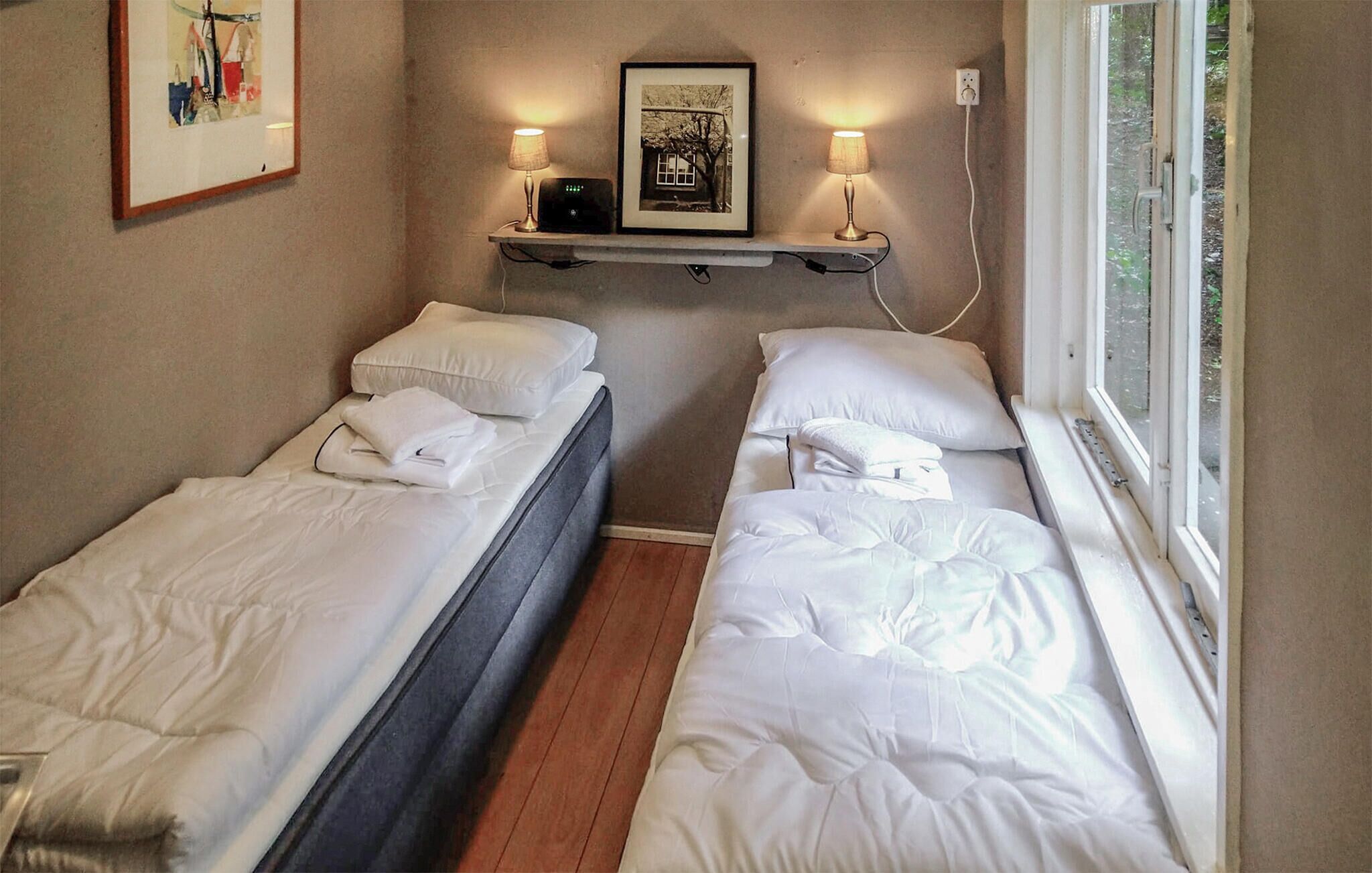 A room with two single beds, a small shelf with a framed picture, and a window with a view of greenery.