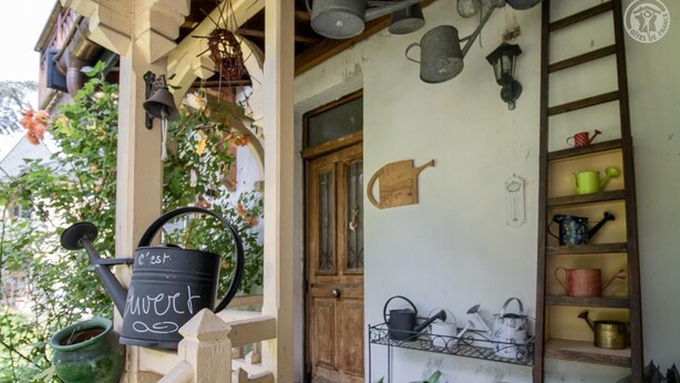A rustic porch with a black watering can labeled 'c'est l'herbe' and various gardening tools on shelves and walls. - Watering Can And Soldier (PLANAY)