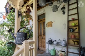 A rustic porch with a black watering can labeled 'c'est l'herbe' and various gardening tools on shelves and walls.