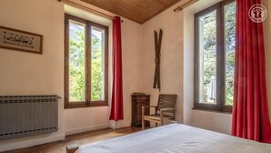 A room with a bed, two windows with red curtains, a chair, and a radiator. - Watering Can And Soldier (PLANAY)
