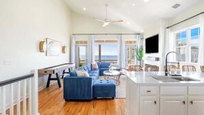 A modern living room with a blue sofa, a dining area with wicker chairs, a kitchen with white cabinets, and a large window offering a view. - Ocean Views-Private Heated Pool & Hot Tub-3 Kings! (Carolina Beach)