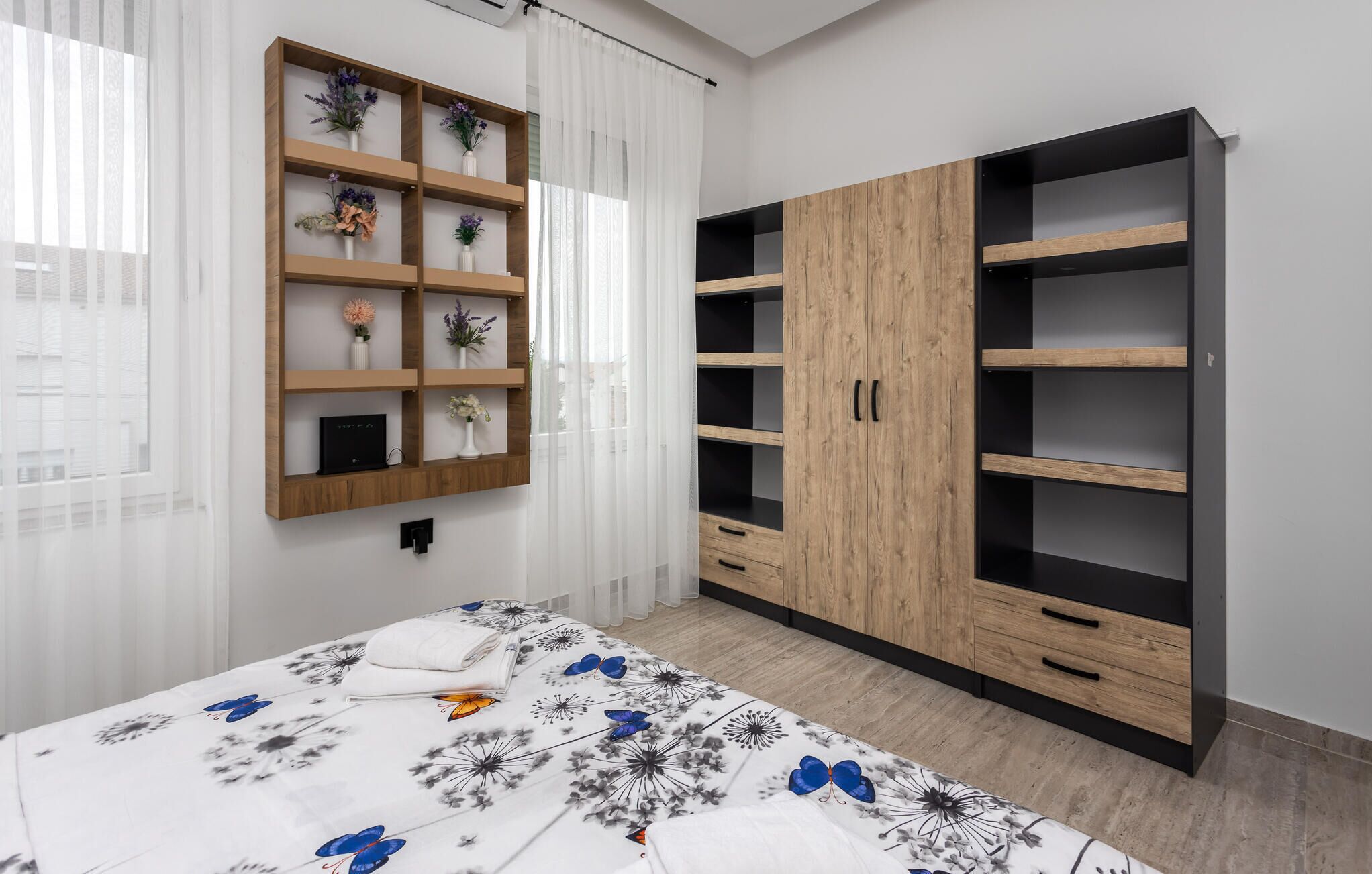A bedroom with a bed, a wooden wardrobe with multiple shelves, and a wall-mounted shelf with decorative items.