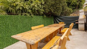 Une table et des bancs en bois, installés à l’extérieur, se dressent sur fond d’une haie bien entretenue et d’une structure en pierre.