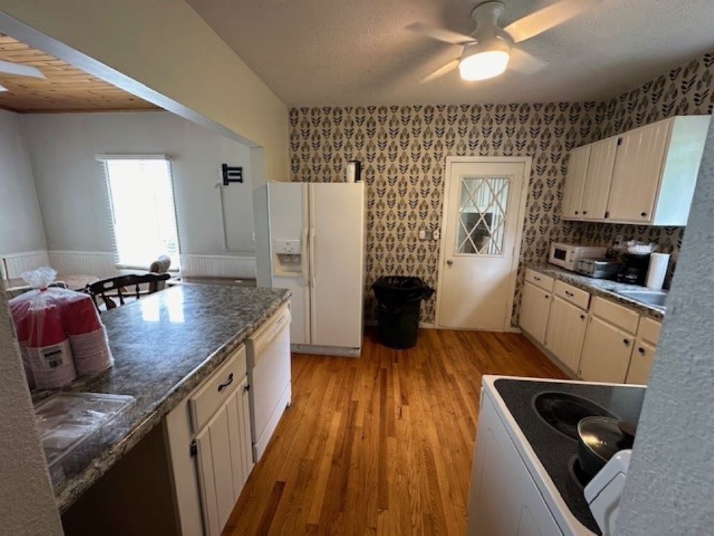A kitchen with wooden flooring, white cabinets, a refrigerator, a microwave, and a trash bin.