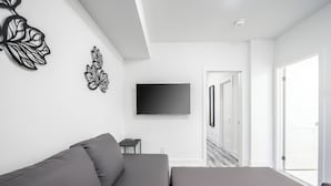 A modern living room with a grey sofa, a flat-screen TV mounted on the wall, and a door leading to another room. - Cozy 1BR with Parking, Only 7 Mins to The Falls and Sleeps 3! (Niagara Falls)