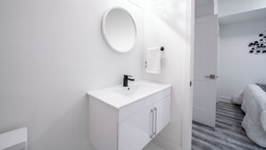 A modern bathroom with a white vanity, a round mirror, and a toilet. - Cozy 1BR with Parking, Only 7 Mins to The Falls and Sleeps 3! (Niagara Falls)