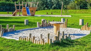 A playground with a wooden slide and climbing structures, surrounded by sand and grassy areas. - Stunning (sta)caravan in Tzummarum (Tzummarum)