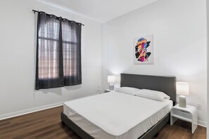 A neatly arranged bedroom with a bed, two bedside tables with lamps, a wall-mounted artwork, and a window with curtains.