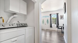 Un área de lavandería con lavadora, secadora y conducto de ventilación. Hay una puerta que da a un comedor con una mesa y sillas.
