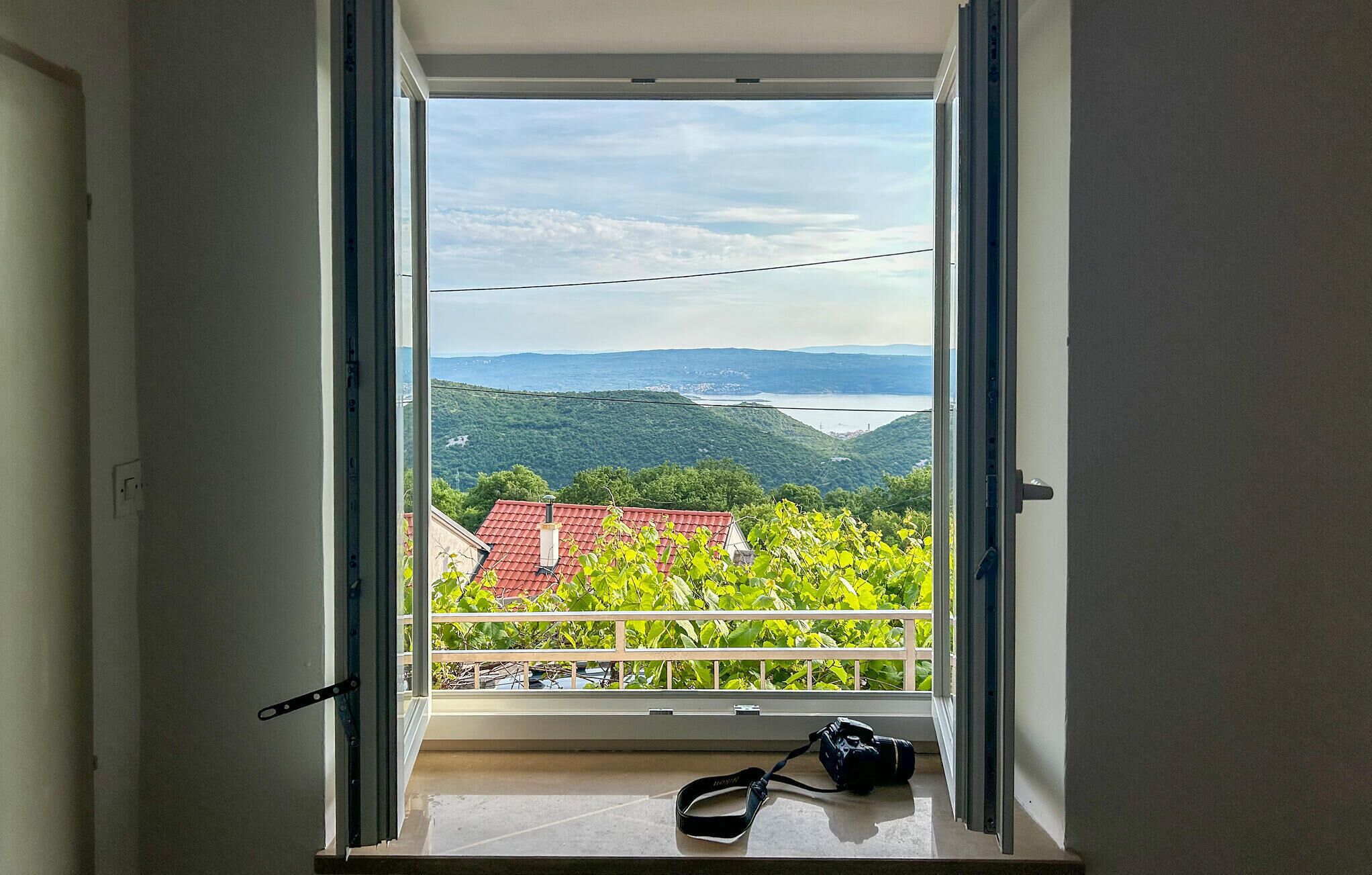 Ein Zimmer mit großem Fenster, einer Kamera auf dem Sims und Blick auf eine grüne Landschaft mit Gebäuden.