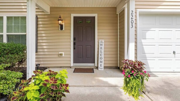 Une maison de couleur beige avec une porte d’entrée marron foncé, une pancarte indiquant « Bienvenue » et un garage portant le numéro 2203.
