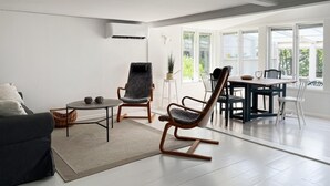 Ein modernes Wohnzimmer mit einer Couch, zwei Schaukelstühlen, einem Esstisch mit Stühlen und einem großen Fenster, das viel Tageslicht hereinlässt.